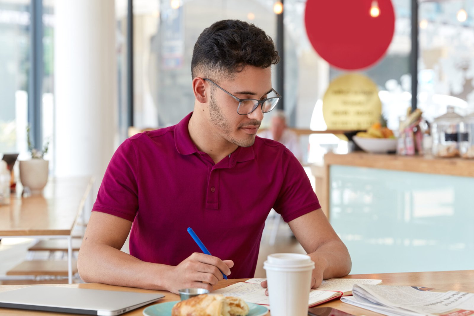 Homem sentado em um café faz anotações em um caderno enquanto trabalha no laptop, representando a capacitação contínua como parte da rotina de aprendizado.