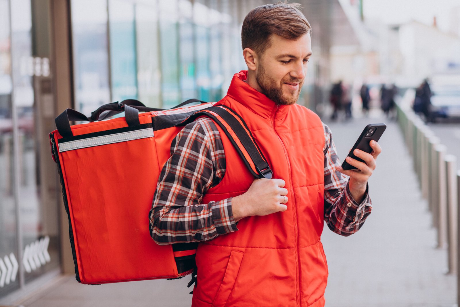 Entregador de delivery com mochila térmica vermelha olhando para o celular, representando soluções para aumentar vendas no delivery de restaurantes.